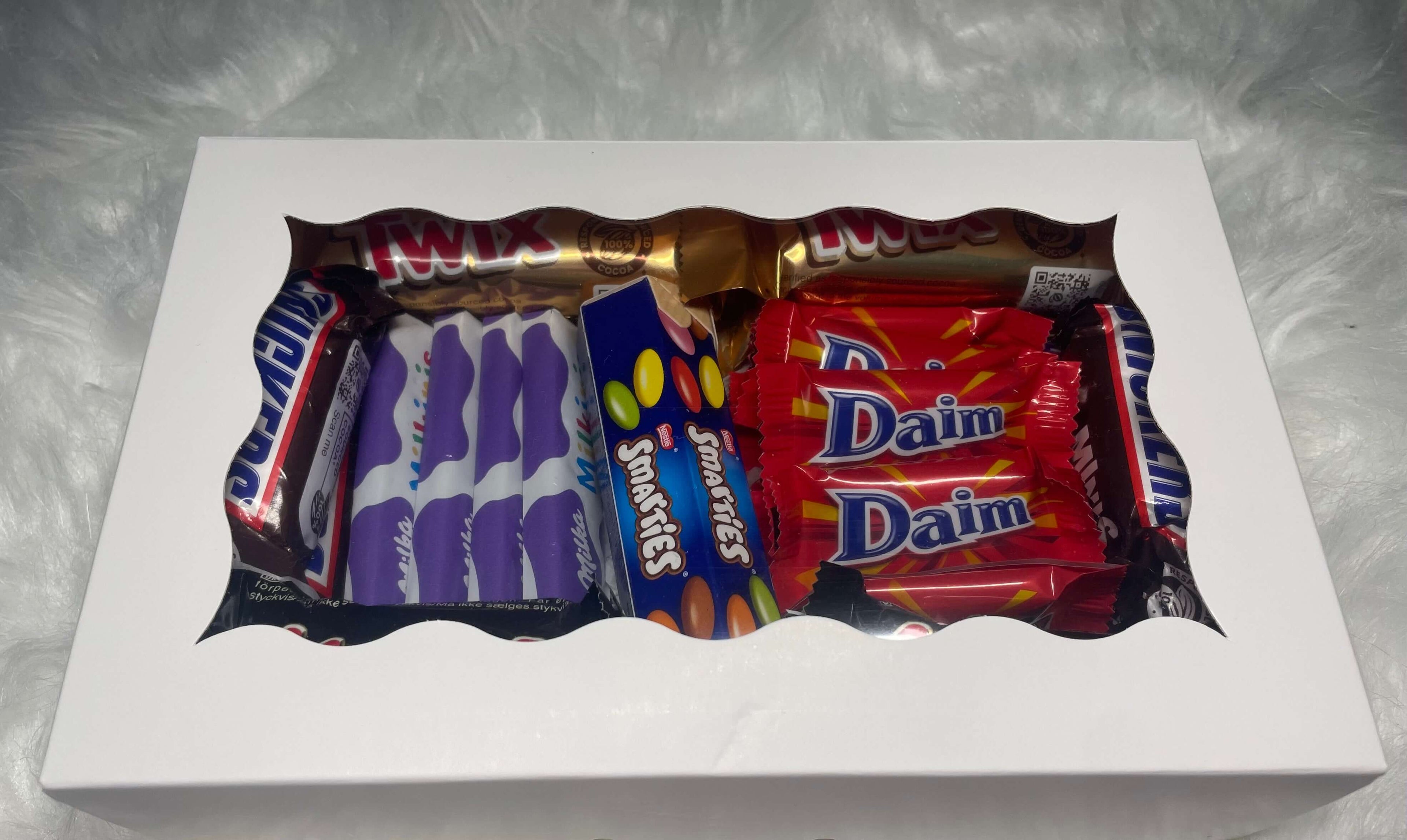 Coffret cadeau blanc fermé avec fenêtre transparente montrant un assortiment de chocolats : Twix, Snickers, Mars, Milka, Daim et Smartie.
